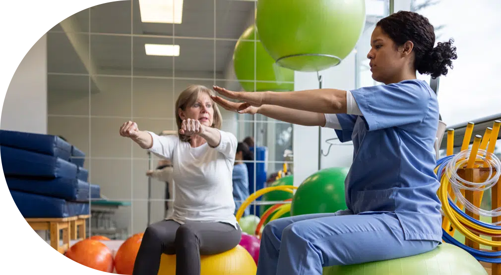 Young woman nurse in scrubs sitting on a yoga ball and holding her arms out to instruct her patient, to the left of her, who is doing the same movement.