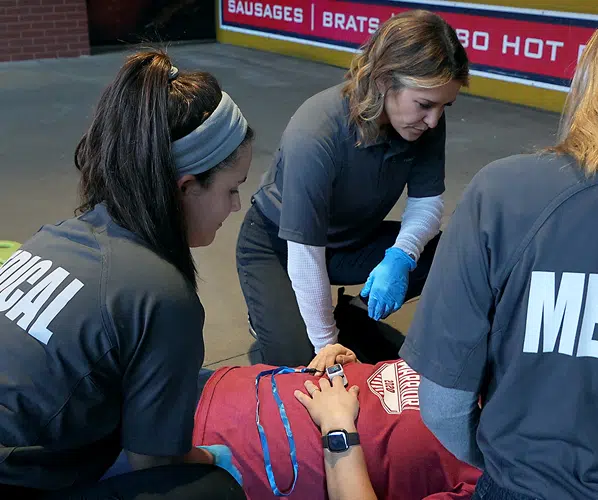Emergency medical team providing on-site care to an injured person wearing a red shirt. The responders, dressed in gray 'MEDICAL' uniforms and gloves, assess the patient's condition in a public venue