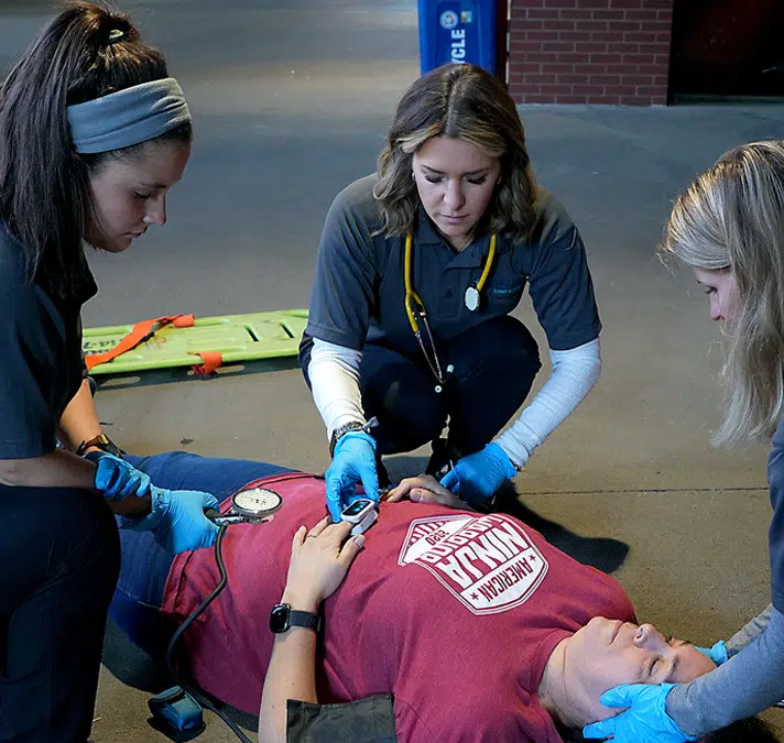 Emergency medical team providing on-site care to an injured person wearing a red shirt. The responders, dressed in gray 'MEDICAL' uniforms and gloves, assess the patient's condition in a public venue